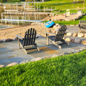 Photo of two chairs on a brick paver patio surrounded by small boulders and green grass with a beach in and lake in the back