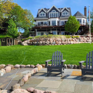 Photo of a large residential house with a retaining wall made out of boulders and chairs on a brick paver patio in the foreground